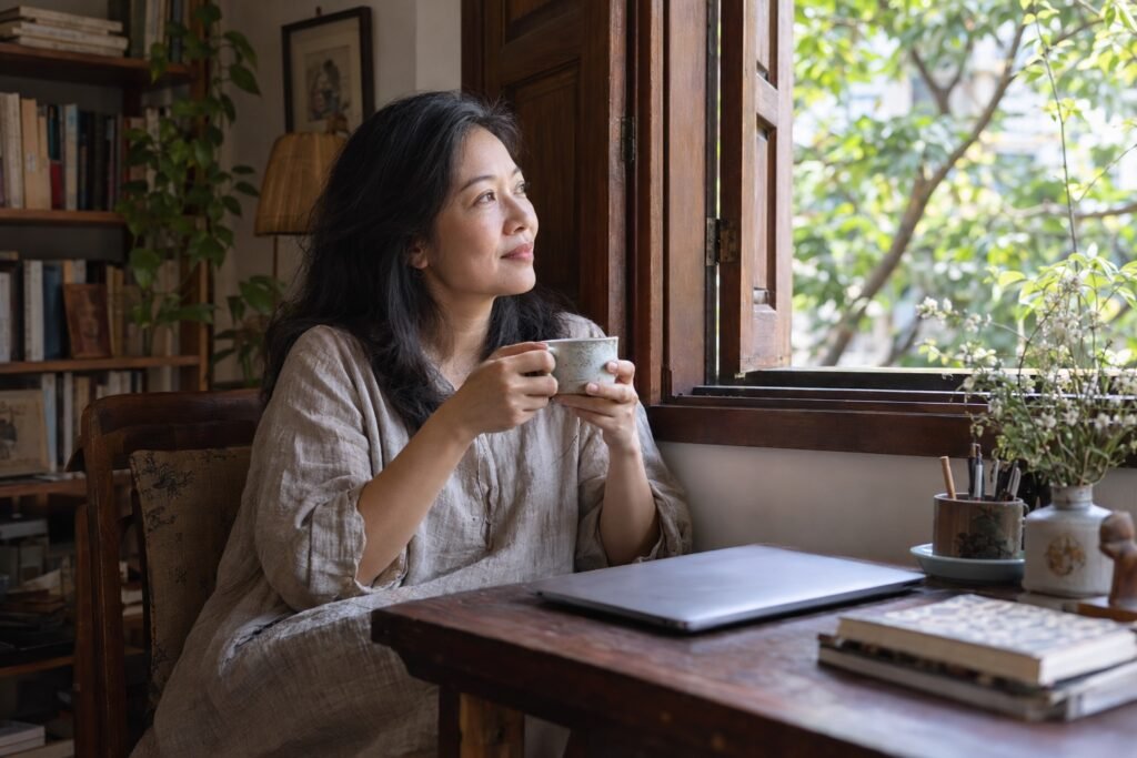 Người phụ nữ ngồi bên cửa sổ gỗ, cầm tách trà, máy tính gập lại, nhìn ra tán lá